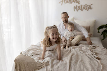 Porträt eines glücklichen kleinen Mädchens auf dem Bett zu Hause mit Vater und Bruder, die sie aus dem Hintergrund beobachten - GMLF00117