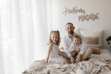 Familienporträt eines glücklichen Vaters und seiner beiden Kinder, die zusammen auf einem Bett sitzen - GMLF00118