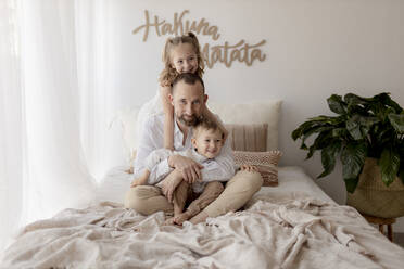 Family portrait of happy father sitting on bed with his two children - GMLF00119