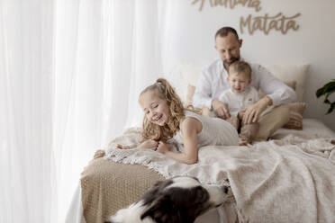 Porträt eines lächelnden kleinen Mädchens auf einem Bett mit Vater und Bruder im Hintergrund - GMLF00136