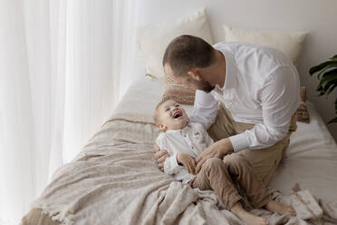 Father crouching on bed tickling his little son - GMLF00153