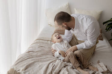 Vater hockt auf dem Bett und kitzelt seinen kleinen Sohn - GMLF00153