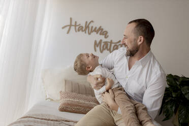 Father crouching on bed tickling his little son - GMLF00157