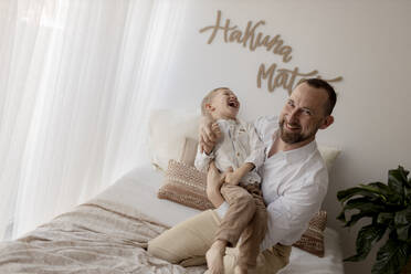 Father crouching on bed tickling his little son - GMLF00158