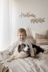 Porträt eines glücklichen kleinen Jungen auf dem Bett mit seinem Hund - GMLF00161