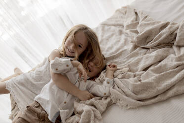 Porträt eines kleinen Mädchens, das seinen lachenden Bruder auf dem Bett kuschelt - GMLF00174