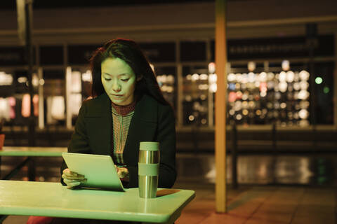 Junge Frau mit Tablet auf der Terrasse eines Cafés in der Stadt bei Nacht, Frankfurt, Deutschland, lizenzfreies Stockfoto