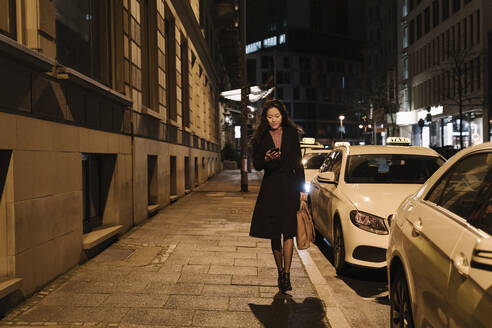 Junge Frau, die nachts in der Stadt spazieren geht und ihr Smartphone benutzt, Frankfurt, Deutschland - AHSF02416