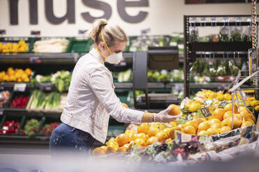 Teenage girl wearing protectice mask and gloves choosing fruits at supermarket - ASCF01254