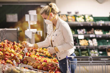 Jugendliches Mädchen mit Schutzmaske und Handschuhen beim Aussuchen von Äpfeln im Supermarkt - ASCF01256