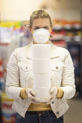 Teenage girl wearing protectice mask and gloves holding stack of four toilet rolls at supermarket - ASCF01263