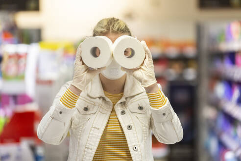 Teenager-Mädchen mit Schutzmaske und Handschuhen, das durch die Löcher von Toilettenrollen im Supermarkt schaut - ASCF01264