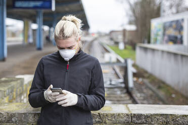 Teenage girl wearing protective mask and gloves using cell phone while waiting at train station - ASCF01266