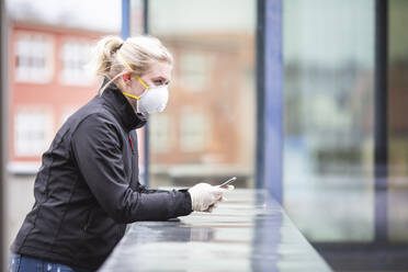 Blond teenage girl with smartphone wearing protective mask and gloves using outdoors - ASCF01267