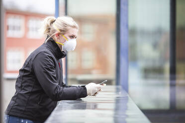 Blondes Teenager-Mädchen mit Smartphone, das Schutzmaske und Handschuhe trägt und im Freien arbeitet - ASCF01267