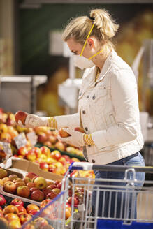 Jugendliches Mädchen mit Schutzmaske und Handschuhen beim Aussuchen von Äpfeln im Supermarkt - ASCF01272