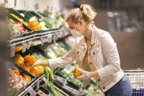 Teenagerin mit Schutzmaske und Handschuhen beim Aussuchen von Paprika im Supermarkt - ASCF01273