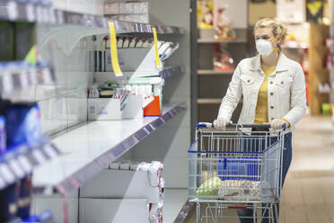 Teenage girl wearing protectice mask and gloves looking at empty shelves at supermarket - ASCF01275