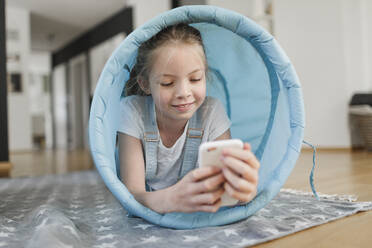 Portrait of smiling girl lying in blue fabric tunnel on the floor at home looking at cell phone - HMEF00921