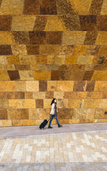 Young businesswoman with trolley bag walking in front of stone wall - XLGF00105