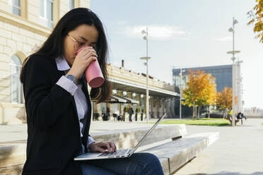 Junge Geschäftsfrau mit wiederverwendbarem Becher sitzt auf einer Bank im Freien und arbeitet an einem Laptop - XLGF00120