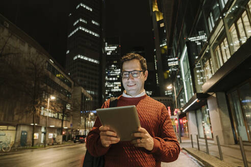 Porträt eines lächelnden Unternehmers in der Stadt mit Blick auf ein digitales Tablet bei Nacht, Frankfurt, Deutschland - AHSF02426