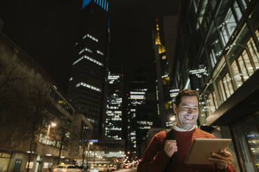 Porträt eines glücklichen Unternehmers in der Stadt mit Blick auf ein digitales Tablet bei Nacht, Frankfurt, Deutschland - AHSF02427