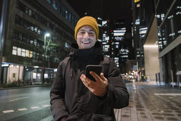 Porträt eines lächelnden Mannes mit Blick auf sein Smartphone bei Nacht, Frankfurt, Deutschland - AHSF02429