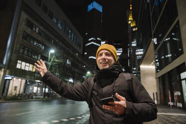 Porträt eines lächelnden Mannes mit Smartphone, der nachts ein Taxi anhält, Frankfurt, Deutschland - AHSF02430