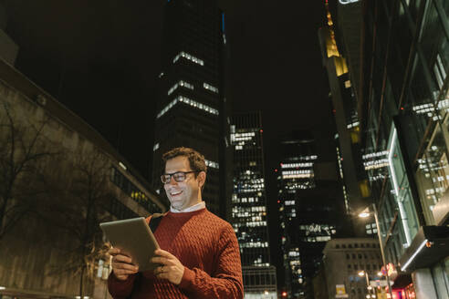 Porträt eines glücklichen Unternehmers in der Stadt mit Blick auf ein digitales Tablet bei Nacht, Frankfurt, Deutschland - AHSF02438
