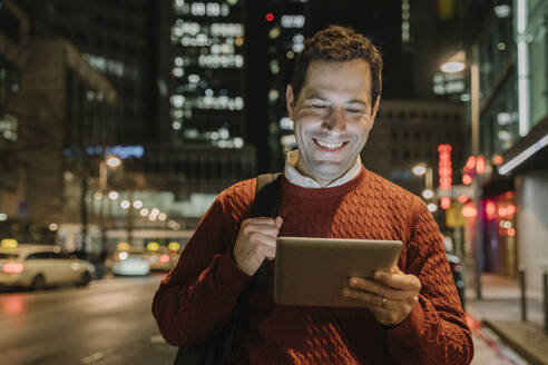 Porträt eines glücklichen Unternehmers in der Stadt mit Blick auf ein digitales Tablet bei Nacht, Frankfurt, Deutschland - AHSF02439