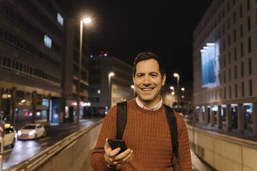 Portrait of smiling businessman with smartphone at night, Frankfurt, Germany - AHSF02445