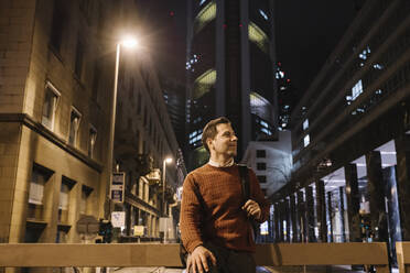 Smiling businessman with backpack in the city at night, Frankfurt, Germany - AHSF02446