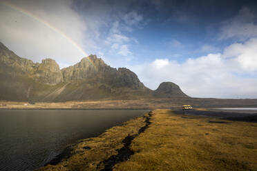 Scenic view of sea amidst mountains against rainbow in sky at Hvalnes Nature Reserve Beach, Iceland - DAMF00422