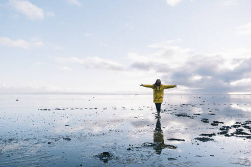 Woman with arms outstretched reflecting in sea at Hvalnes Nature Reserve Beach, Iceland - DAMF00427