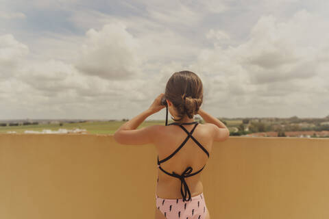 Rückenansicht eines Mädchens im Badeanzug, das auf einer Dachterrasse steht und durch ein Fernglas schaut, lizenzfreies Stockfoto