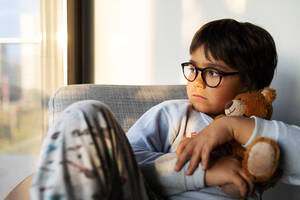 Porträt eines ernsten Jungen mit Teddybär, der zu Hause auf einem Sessel sitzt und aus dem Fenster schaut - VABF02860