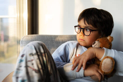 Portrait of serious boy with teddy bear sitting on armchair at home looking out of window - VABF02860