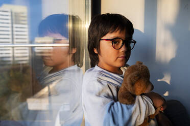 Portrait of serious boy with teddy bear leaning at window in the evening - VABF02861