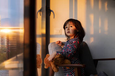 Portrait of little girl with teddy bear looking out of window at sunset - VABF02867