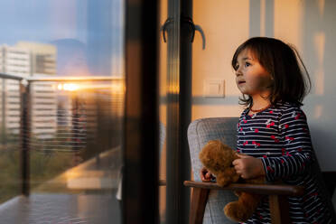 Porträt eines kleinen Mädchens mit Teddybär, das bei Sonnenuntergang aus dem Fenster schaut - VABF02868