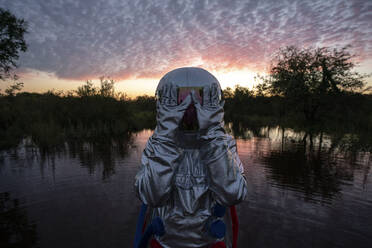 Porträt einer im Wasser stehenden Raumfahrerin bei Sonnenuntergang, die Hände bedecken den Helm - VPIF02405