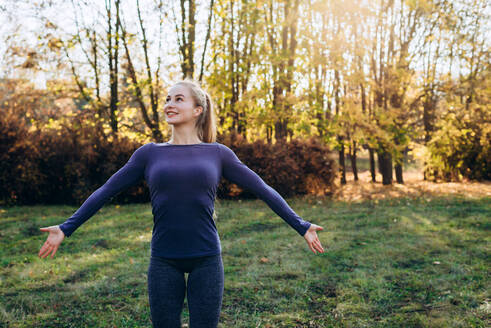 Schönes lächelndes Mädchen bei der Morgengymnastik im Park, das mit ausgebreiteten Armen wegschaut. - CAVF80533