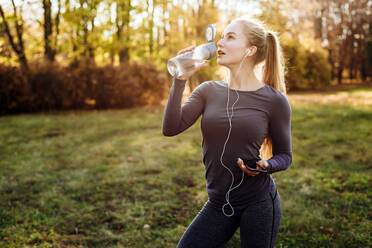 Fitness in the park, girl drinking water, holding smartphone and headphones in her hand. - CAVF80537