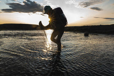 Female Backpacker Crossing Glacial River At Sunset In Highlands - CAVF80650