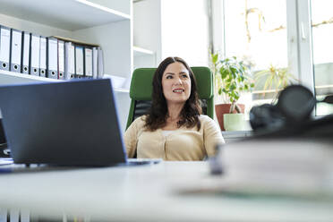Thoughtful businesswoman with laptop sitting at desk while looking away in office - MMIF00206