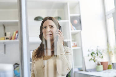 Geschäftsfrau spricht auf Smartphone durch Glas im Büro gesehen - MMIF00207