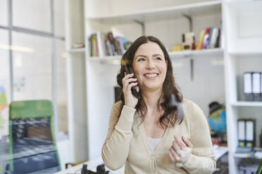 Lächelnde Geschäftsfrau, die mit einem Mobiltelefon spricht, gesehen durch Glas im Büro - MMIF00209