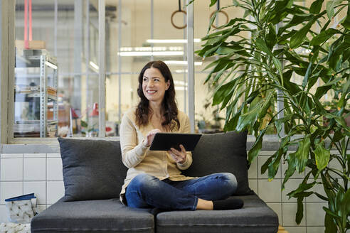 Smiling businesswoman using digital tablet while sitting on sofa at office - MMIF00213