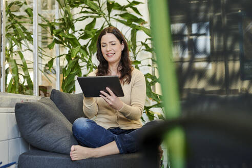 Lächelnder Unternehmer, der ein digitales Tablet benutzt, während er auf dem Sofa am Arbeitsplatz sitzt - MMIF00214